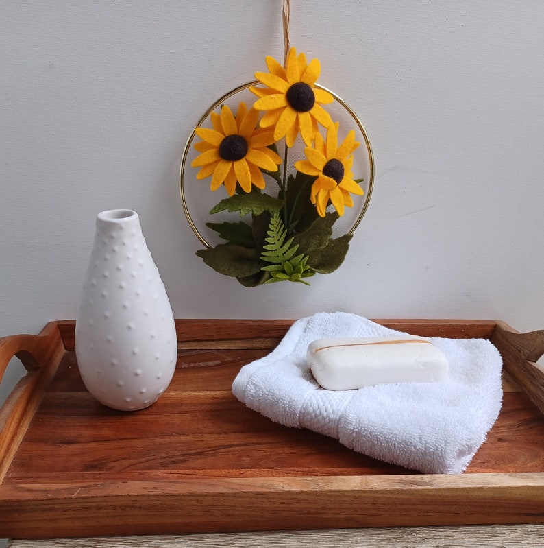 Puede incluir: Una bandeja de madera con una toalla blanca, un jarr&oacute;n de cer&aacute;mica blanca y un aro de metal dorado con tres girasoles de fieltro.