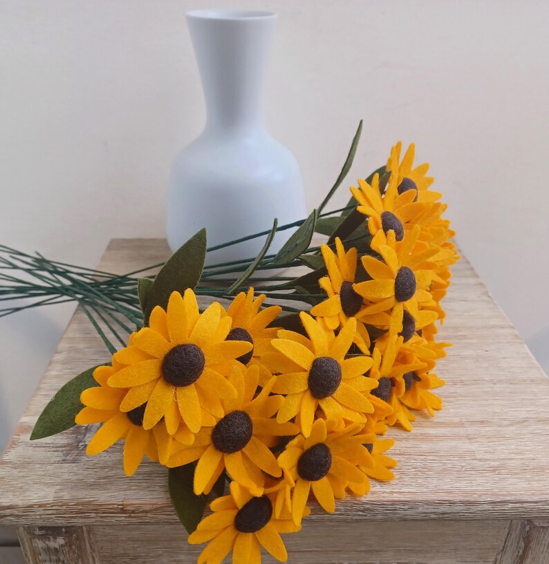 Op de afbeelding: Een boeket van viltzonnebloemen in tinten geel en bruin. De bloemen zijn in een vaas gerangschikt en hebben groene stelen.