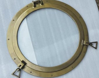 Könnte beinhalten: Rundes Bullauge aus Messing mit klarer Glasscheibe in der Mitte. Das Bullauge hat ein Scharnier oben und zwei seitliche Verschlüsse. Der Rahmen ist mit kleinen Nieten befestigt, das Design erinnert an nautische Dekoration. Durchmesser ca. 30 cm.