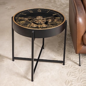 Pode incluir: Uma mesa lateral redonda preta com um design de mostrador de relógio. O tampo da mesa apresenta um fundo preto com ponteiros, engrenagens e algarismos romanos dourados. A mesa tem uma moldura dourada e pernas de metal preto. A mesa tem cerca de 61 cm de diâmetro.