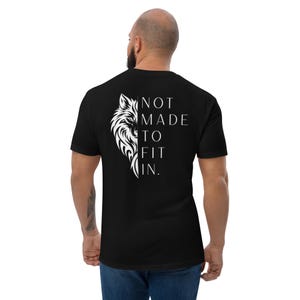 May include: A black short-sleeved t-shirt featuring a white graphic on the back. The design displays the left half of a wolf's face next to the text "NOT MADE TO FIT IN." printed in a clean, sans-serif font. The shirt has a crew neckline.
