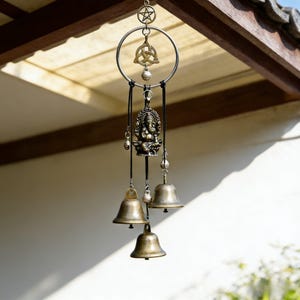 May include: A bronze-toned wind chime with a circular frame, featuring a pentagram, a Celtic knot, and a detailed figure of a deity. The chime includes several small bells suspended by black cords, creating a decorative and spiritual piece.
