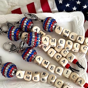 May include: Keychains with red, white, and blue rhinestone-covered spheres and wooden letter beads spelling "AMERICA." The keychains have silver-colored clasps. The background includes an American flag.