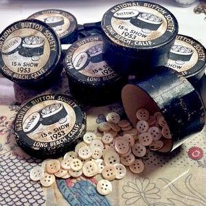 May include: Several round black cardboard containers labeled National Button Society 15th Show 1953 Long Beach Calif. One container is tipped over, spilling small, round, four-hole cream-colored buttons onto the surface. The lids feature a graphic illustration.
