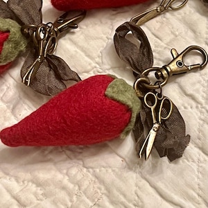 May include: Close-up of several red felt chili pepper keychains with green stems. Each keychain has a bronze-colored clasp and a small pair of scissors charm, attached to a brown ribbon.