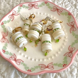 May include: A collection of white button charms with gold-colored clasps and green rhinestone accents. Each charm is adorned with a small green bead and a piece of measuring tape ribbon. The charms are arranged on a decorative plate with a pink bow and berry design.