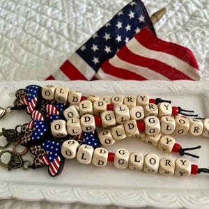 May include: Keychains with wooden lettered beads spelling "OLD GLORY" are displayed on a white tray. The keychains have red and black accents. Small American flags are in the background, with a white background.