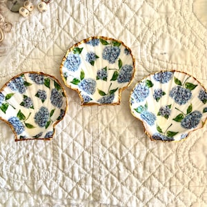 May include: Three seashell-shaped dishes with a white background, adorned with blue hydrangea floral patterns and green leaves. The edges are trimmed with gold. The dishes are arranged on a white quilted surface.