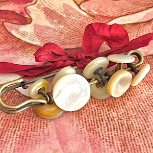 May include: A brass safety pin brooch adorned with multiple round, off-white mother-of-pearl buttons of varying sizes attached to the metal bar, accented with a tied dark red ribbon bow secured at the center of the pin mechanism.
