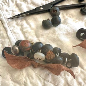 May include: An assortment of vintage buttons in various shapes, sizes, and colors, including black, silver, and white. A rusty-colored ribbon and black scissors are also present, all arranged on a white, quilted surface.