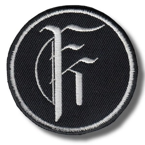 Op de afbeelding: Een ronde, zwarte geborduurde patch met een witte rand. De patch toont een gestileerde witte letter "F" in een gotisch lettertype, gecentreerd. Het ontwerp heeft een klassieke, vintage esthetiek, geschikt voor kleding of accessoires.