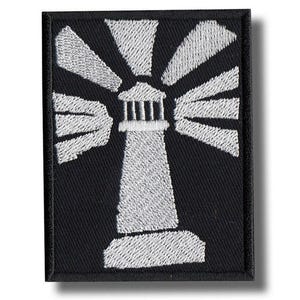 May include: Black embroidered patch featuring a white lighthouse with radiating beams of light. The lighthouse design is centered on a black rectangular background. The patch is likely for clothing or accessories.