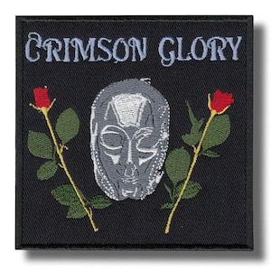 May include: Square black patch with the words "CRIMSON GLORY" embroidered in light blue. The patch features a gray mask design and two red roses with green leaves and stems.
