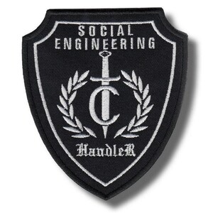 Peut inclure: Écusson brodé noir et blanc en forme de bouclier. Le patch comporte les mots "SOCIAL ENGINEERING Handler" en blanc, avec une épée et une couronne de laurier. Le patch est probablement destiné à une veste ou un sac.