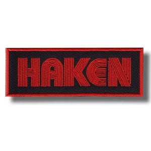 Puede incluir: Parche bordado rectangular con la palabra "HAKEN" en rojo. El texto es estilizado y se encuentra sobre un fondo negro con un borde rojo. El parche es probablemente de tela.