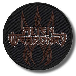 May include: A circular black patch featuring the words "ALIEN WEAPONRY" in a brown, textured font with white outlines. Brown flame-like designs are above and below the text. The patch has a black border and is suitable for clothing.