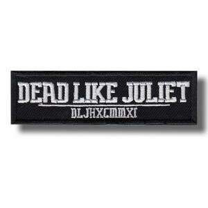 Puede incluir: Un parche rectangular negro con texto bordado en blanco que dice "DEAD LIKE JULIET" encima de "DLJHXCMMXI". El parche es probablemente para ropa o accesorios, con una fuente audaz de estilo gótico.