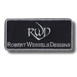 Puede incluir: Un parche rectangular negro con un borde gris. El parche presenta el texto bordado "ROBERT WESSELS DESIGNS" en blanco, junto con un logotipo "RWD" estilizado en blanco. El parche es probablemente para ropa o accesorios.