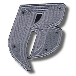Puede incluir: Un parche bordado gris con la forma de la letra "B". El parche tiene un aspecto texturizado con una serie de líneas paralelas y pequeños remaches oscuros. La letra está delineada en un gris más oscuro.