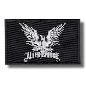Op de afbeelding: Een rechthoekige zwarte patch met een wit geborduurd ontwerp. Het ontwerp toont een gestileerde vogel met uitgespreide vleugels boven de tekst "ALTER BRIDGE" in een decoratief lettertype. De patch heeft een zwarte rand.