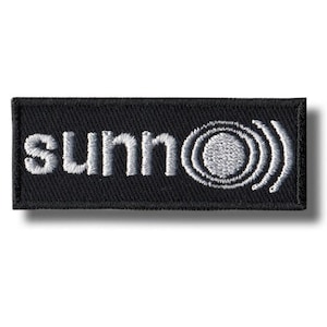 May include: Embroidered patch with the word "sunn" in white, stylized text on a black rectangular background. The design includes a circular graphic with radiating lines, suggesting sound waves or a similar concept.