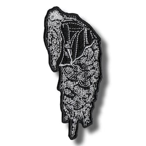 May include: Black and white embroidered patch featuring a stylized angel wing design. The patch has a detailed, textured appearance with intricate stitching. The wing shape is elongated and slightly curved, with a black border.