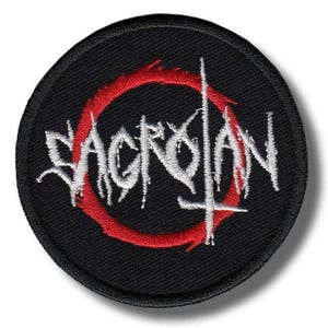 Könnte beinhalten: Ein runder, schwarzer Aufnäher mit dem Wort "SACROLAN" in weißer Stickerei, umgeben von einem roten Kreis und einem kreuzartigen Design. Geeignet für Kleidung oder Accessoires.