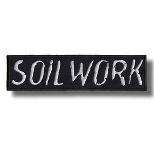 May include: A black rectangular patch with the word "SOIL WORK" embroidered in white, stylized lettering. The patch has a black border and is likely designed to be sewn or ironed onto clothing or accessories.