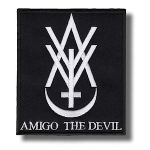 May include: Black embroidered patch featuring a white stylized design with a cross, crescent, and geometric shapes. The text "AMIGO THE DEVIL" is embroidered in white at the bottom of the patch. The patch is square-shaped.