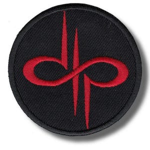 Puede incluir: Un parche redondo negro con un diseño rojo bordado. El diseño presenta un símbolo de infinito estilizado con dos líneas verticales. El parche tiene un borde cosido, probablemente un logotipo.