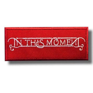 Peut inclure: Un écusson rectangulaire rouge avec le texte brodé "IN THIS MOMENT" en blanc, orné de volutes décoratives. Le patch a un aspect texturé et une ombre subtile.