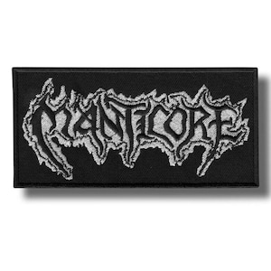 Peut inclure: Écusson rectangulaire noir avec le mot "MANTCORE" brodé en blanc, dans une police gothique stylisée. Les lettres sont bordées d'un motif en forme de flamme. Pour vêtements ou accessoires.