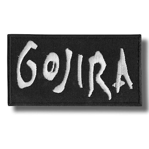Könnte beinhalten: Schwarzer rechteckiger Aufnäher mit dem Wort "GOJIRA" in Weiß gestickt. Die Buchstaben sind stilisiert und scheinen in einer fetten, leicht verblassten Schriftart zu sein. Der Aufnäher hat einen schwarzen Rand und einen Schatten.