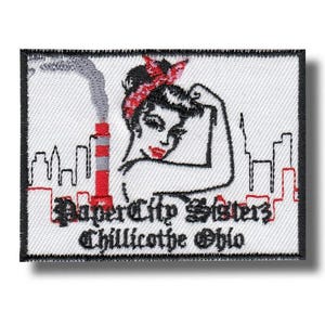 Pode incluir: Patch bordado com uma mulher flexionando o braço, usando uma bandana vermelha e batom. O patch inclui o texto "Paper City Sisters Chillicothe Ohio" e um horizonte com uma chaminé vermelha e cinza.