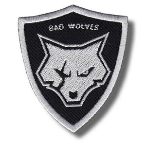 May include: Embroidered patch in a shield shape with a black background and a white wolf head design. The text "BAD WOLVES" is arched across the top. The patch has a white border.