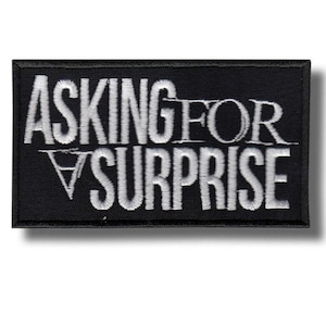 Könnte beinhalten: Schwarzer rechteckiger Aufnäher mit weiß gesticktem Text: „ASKING FOR A SURPRISE“. Der Aufnäher hat einen schwarzen Rand und ist zum Aufnähen oder Aufbügeln auf Kleidung oder Accessoires gedacht.
