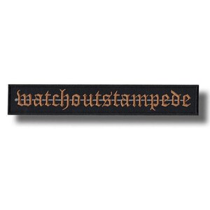 Puede incluir: Un parche rectangular negro con el texto bordado "watchoutstampede" en una fuente gótica. La letra es de color marrón dorado, y el parche tiene un diseño simple y limpio.