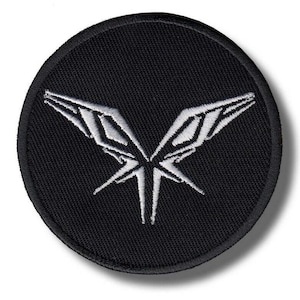Puede incluir: Un parche redondo bordado en negro con un diseño blanco estilizado que se asemeja a una mariposa o alas abstractas. El diseño está centrado en el parche, con costuras detalladas que crean la imagen. El parche tiene un borde negro.