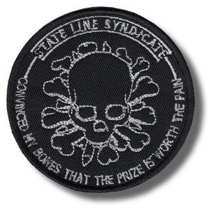 May include: Black, circular patch with white embroidered skull and crossbones design. The text "STATE LINE SYNDICATE" arches around the top, with "CONVINCED MY BONES THAT THE PRIZE IS WORTH THE PAIN" around the bottom.