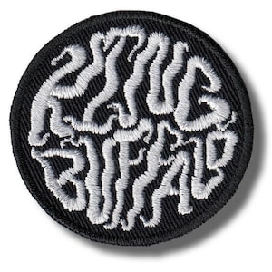 以下が含まれることがあります： 白の「ESSAYS BRAIN」の文字が刺繍された、丸い黒色のパッチ。パッチは、白い糸が黒い背景に対して盛り上がった効果を生み出し、テクスチャード加工された外観になっています。デザインはパッチの中央に配置されています。