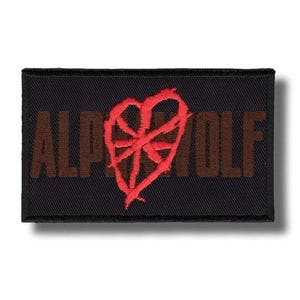 May include: A black rectangular patch featuring the brown text "ALPHA WOLF". A red heart-shaped emblem is embroidered above the text. This patch is designed for application on clothing or accessories.