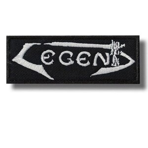 以下が含まれることがあります： 黒い長方形のパッチ。白い刺繍で「LEGEND」の文字が施されています。テキストは装飾的なフォントで、白い矢印のようなデザインで囲まれています。衣類やアクセサリーに適しています。