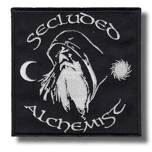 Op de afbeelding: Zwart vierkantige patch met wit geborduurd ontwerp. Het ontwerp toont een figuur met een capuchon en lange baard, een halve maan en een zon. De woorden "Secluded Alchemist" staan in een boog boven en onder de figuur.