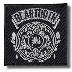 May include: A black square patch embroidered with the word "BEARTOOTH" in white, arched across the top. Beneath, a circular design with an ornate white "B" at the centre, surrounded by detailed floral patterns. The patch has a black border.