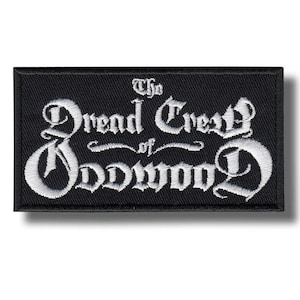 Może przedstawiać: Czarna prostokątna naszywka z białym haftowanym napisem: "The Dread Crest of Oddwood" gotycką czcionką. Naszywka ma czarną obwódkę i jest prawdopodobnie wykonana z tkaniny.