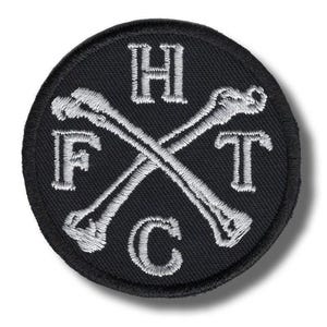 May include: A round, black embroidered patch with white text and crossed bones. The letters "H", "F", "T", and "C" are embroidered around the crossed bones. The patch has a black border.