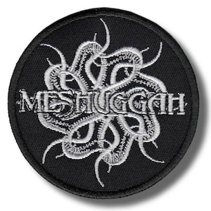 Op de afbeelding: Een ronde, zwarte patch met het woord "MESHUGGAH" in het wit geborduurd, omringd door een wit, gestileerd tentakelontwerp. De patch heeft een zwarte rand en is waarschijnlijk voor kleding of accessoires.
