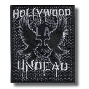 Puede incluir: Parche bordado en negro y gris con las palabras "HOLLYWOOD UNDEAD" y un emblema central con dos aves estilizadas, las letras "LA" y el número "2018". El parche tiene un fondo texturizado.