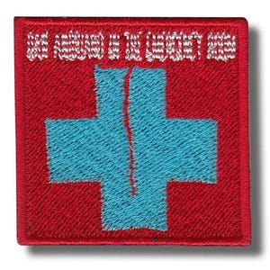 May include: Square red embroidered patch featuring a light blue cross design, bisected by a vertical line. White text is embroidered at the top. The patch has a red border. The design is reminiscent of a medical symbol.