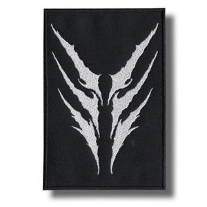 May include: Black rectangular patch featuring a white, symmetrical design, possibly a wolf or abstract creature's face. The design is embroidered on the patch. The patch has a black border.